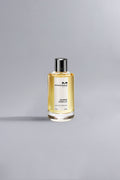 Clear glass perfume bottle with silver cap containing golden liquid, labeled Roses Vanille Ocher, displayed on a light grey background with soft natural lighting.
