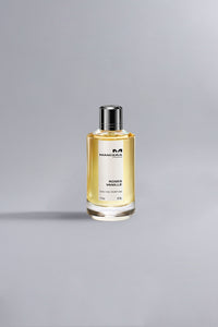 Clear glass perfume bottle with silver cap containing golden liquid, labeled Roses Vanille Ocher, displayed on a light grey background with soft natural lighting.