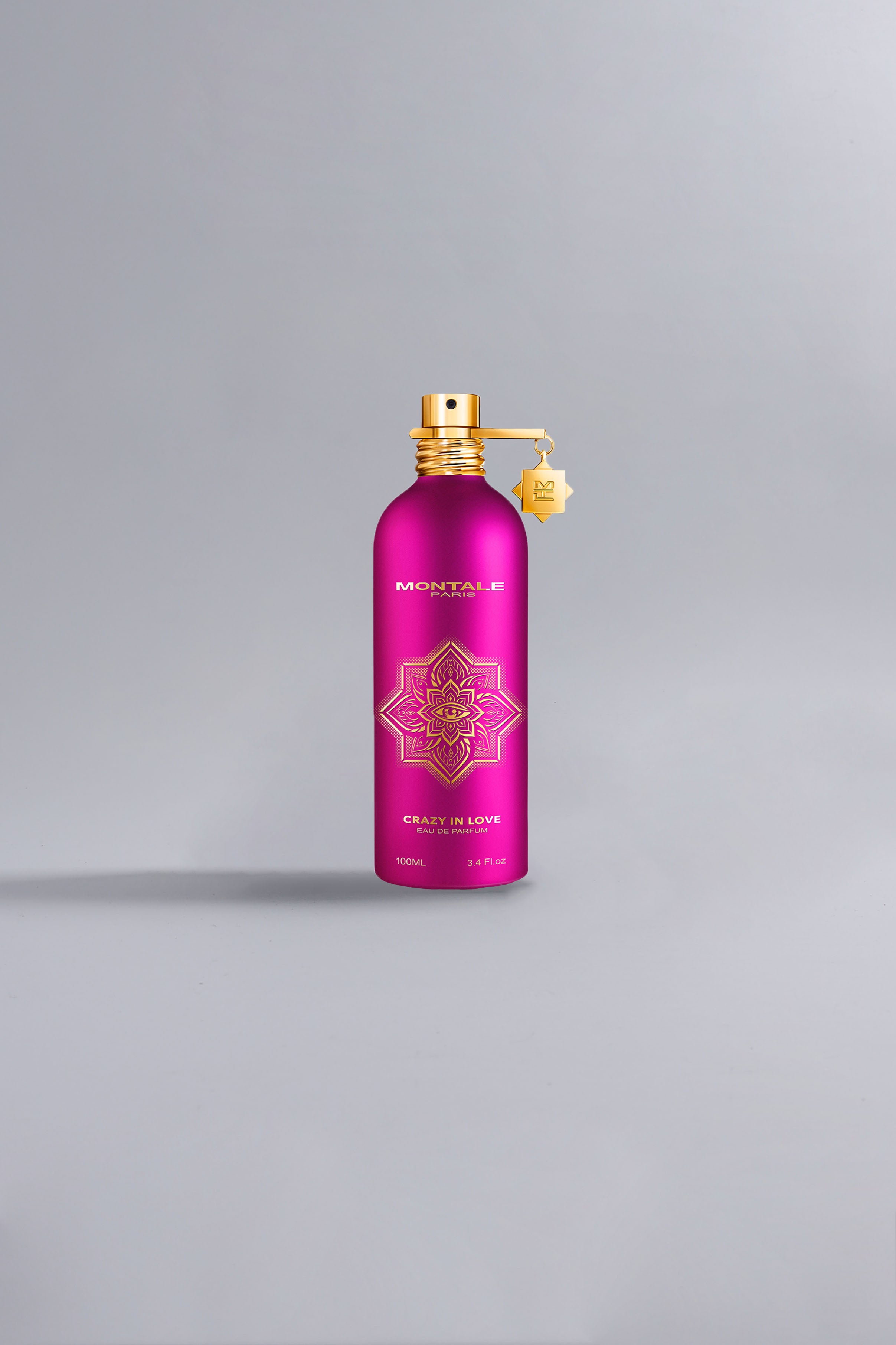 Bright pink luxury perfume bottle with gold intricate design and spray nozzle, labeled Crazy In Love Fuchsia, standing on a smooth neutral background.