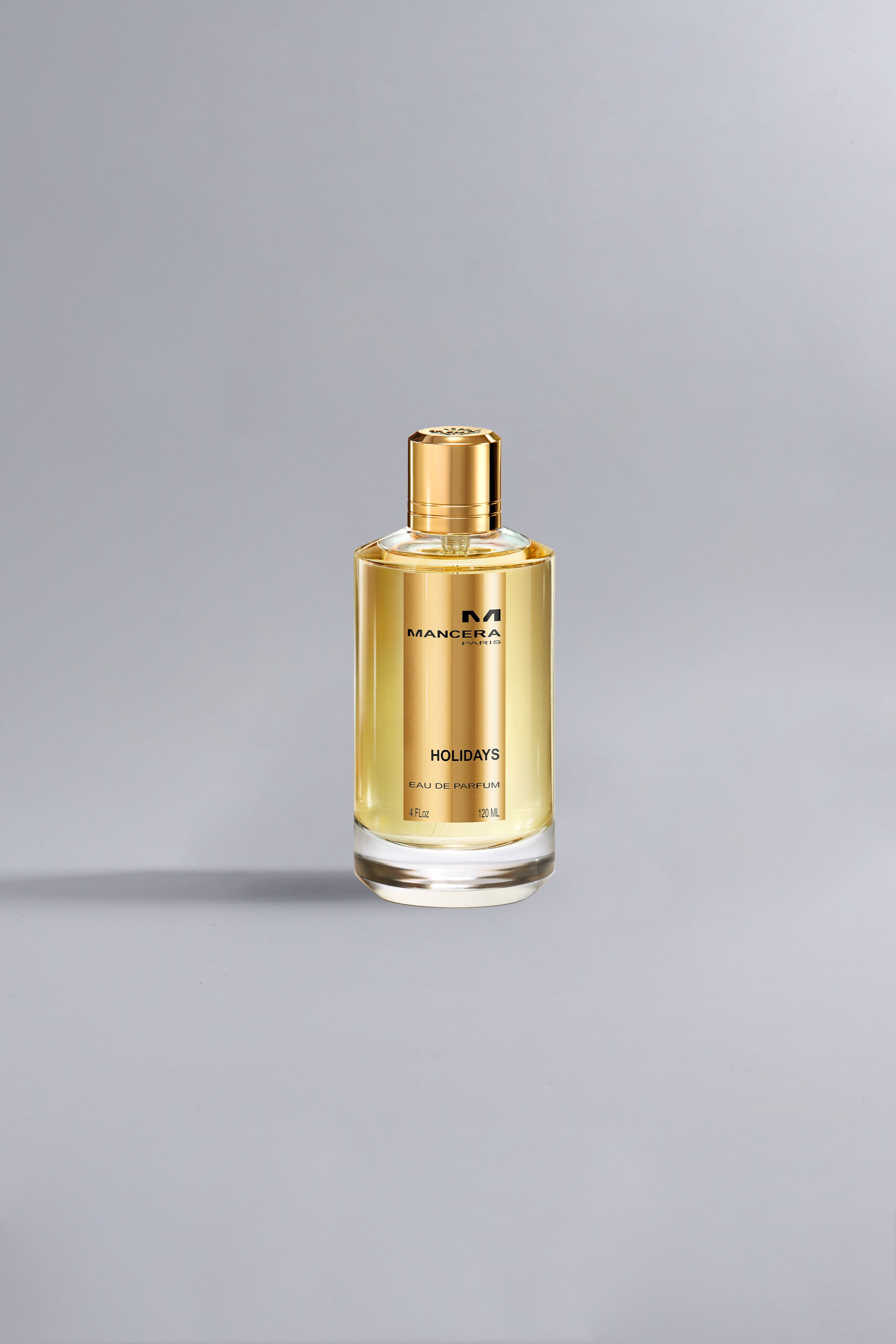 Clear glass bottle of Holidays Gold perfume with golden liquid and gold cap on a neutral gray background, showcasing a luxury fragrance product.