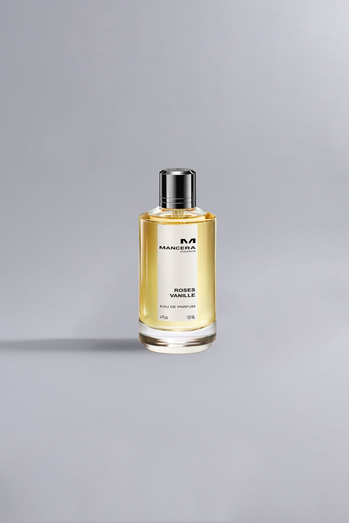 Clear glass perfume bottle with silver cap containing golden liquid, labeled Roses Vanille Ocher, displayed on a light grey background with soft natural lighting.