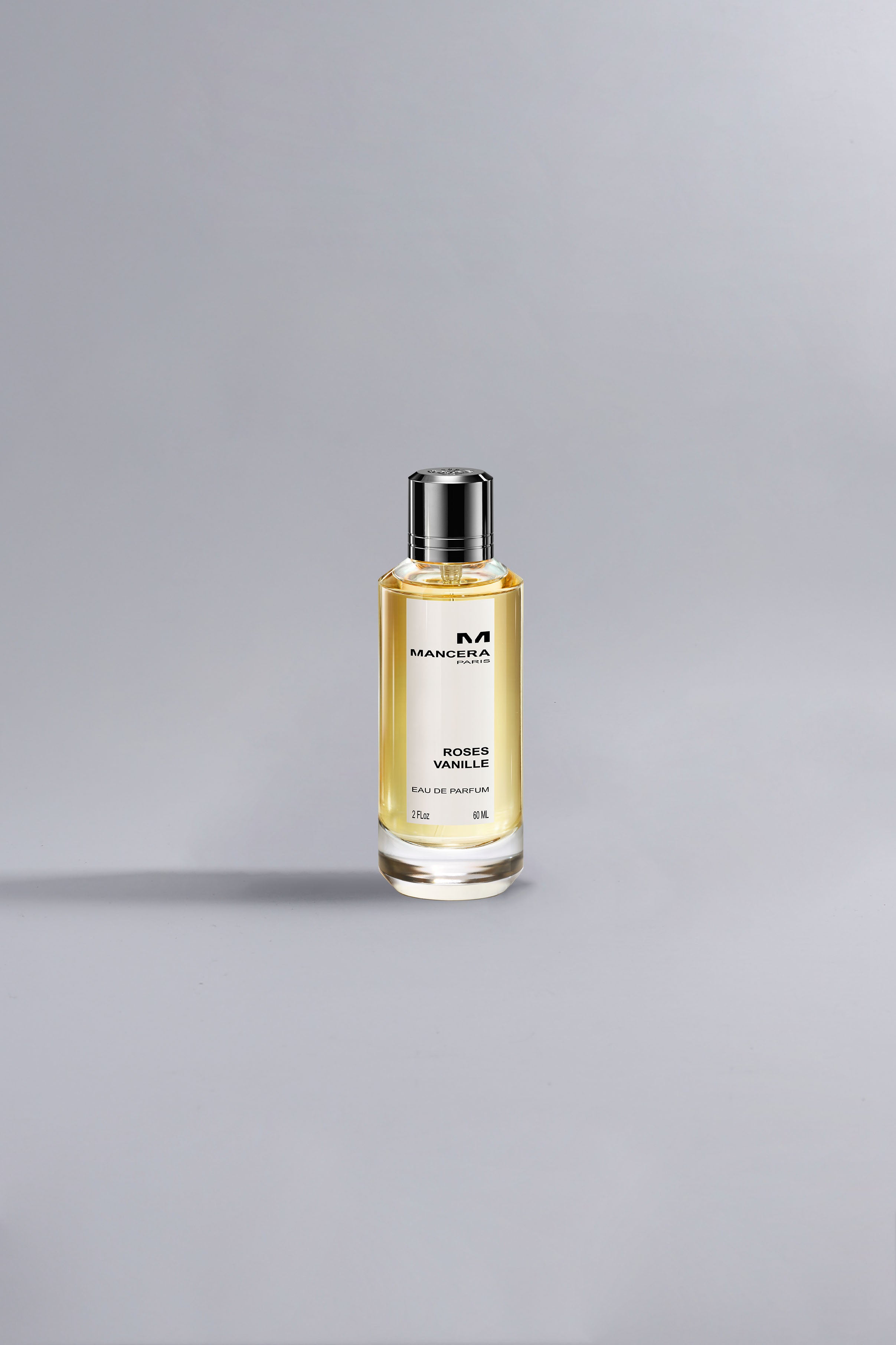 Glass bottle of Roses Vanille Ocher perfume with golden liquid and silver cap, set against a soft gray background, showcasing luxury and elegance.