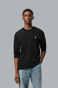 A man wearing a black long-sleeve crewneck shirt with a small white embroidered logo on the left chest, paired with light blue jeans, set against a solid gray background.