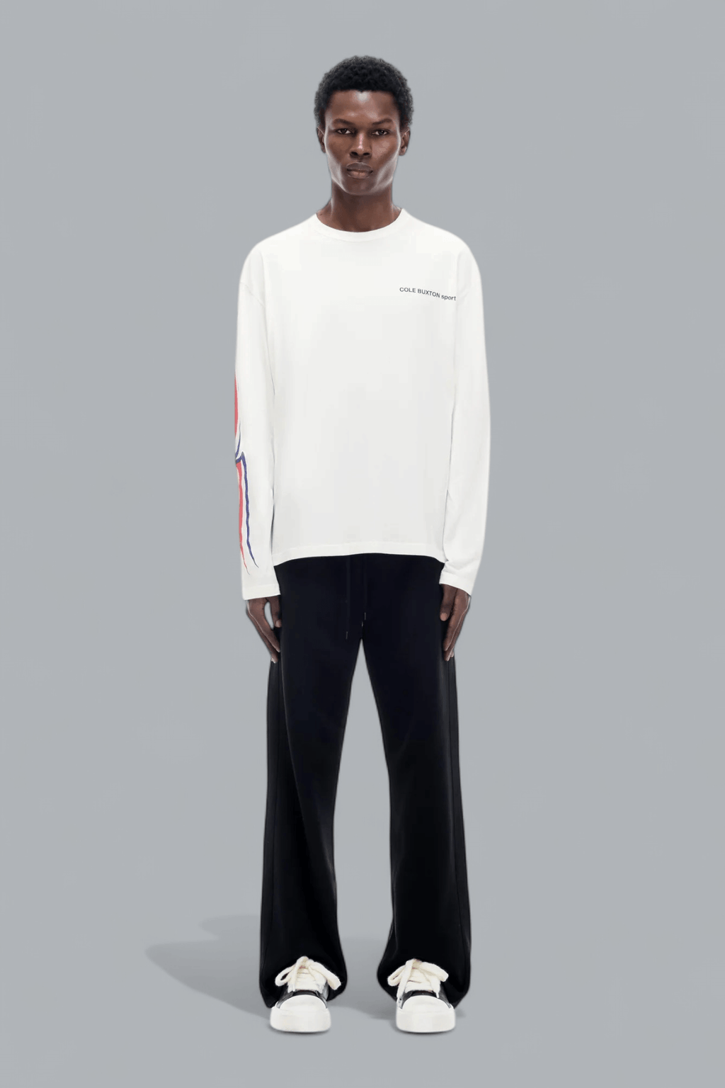 Person wearing a white long-sleeve shirt with colorful accents and black pants on a gray background