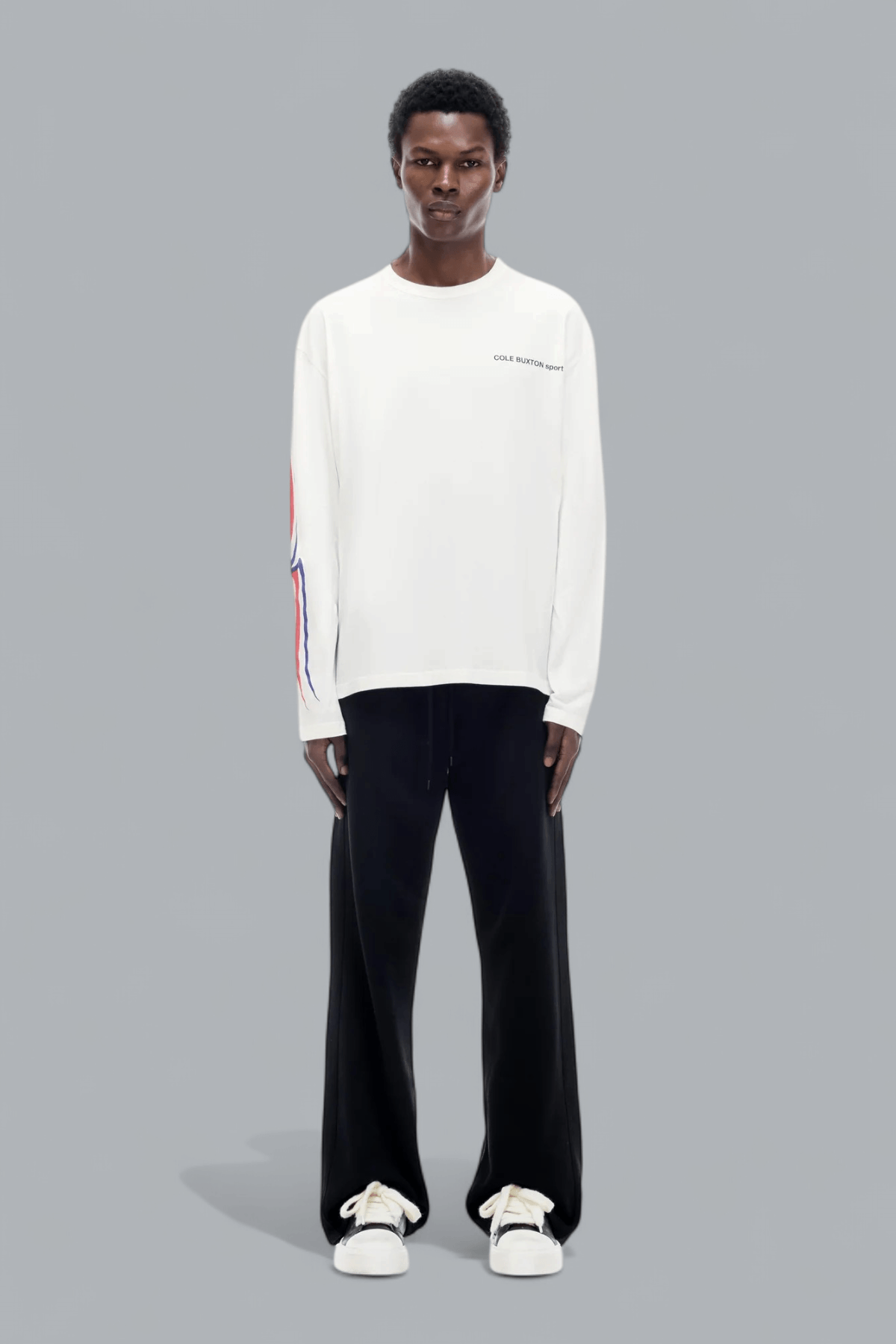 Person wearing a white long-sleeve shirt with colorful accents and black pants on a gray background